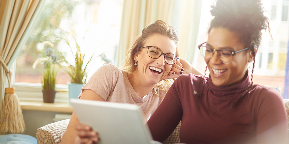 Two women sit on their sofa, online shopping on their digital tablet.