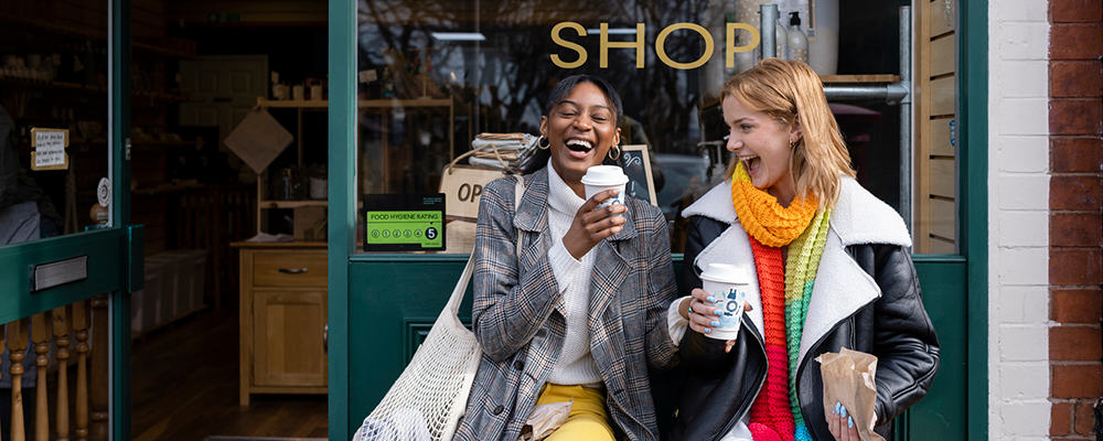 Two friends sitting outside a store, holding coffee and laughing with each other, in the North East of England.